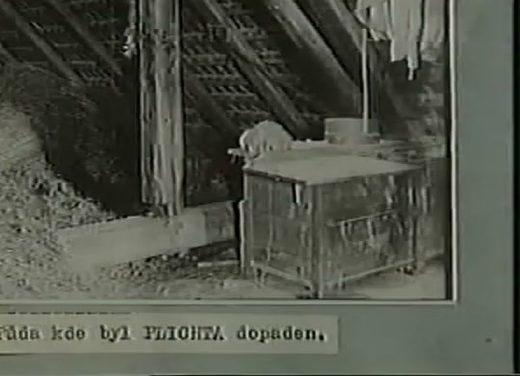 A period photograph of a bare attic under open rafters, a wooden chest beside a plank wall and washing hung from a beam, a typed Czech caption pasted beneath reading the attic where Plichta was caught