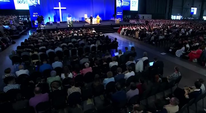 A wide view of the beatification Mass in a Brno exhibition hall, thousands of people seated before a stage with a white cross, the names Jan Bula and Vaclav Drbola under their portraits on the screens above