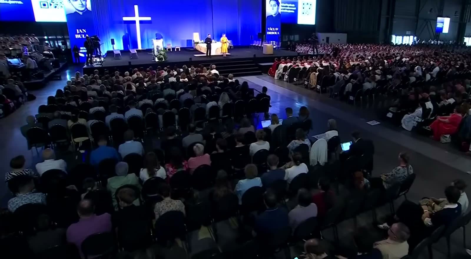 A wide view of the beatification Mass in a Brno exhibition hall, thousands of people seated before a stage with a white cross, the names Jan Bula and Vaclav Drbola under their portraits on the screens above