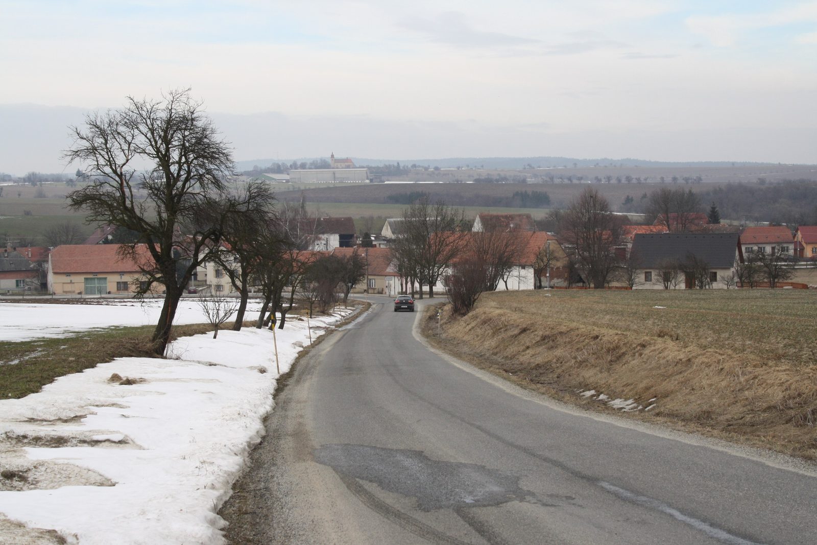 A road running down into the hamlet of Bolikovice from the south, red roofed houses, ploughed fields with patches of snow, a church on the ridge behind