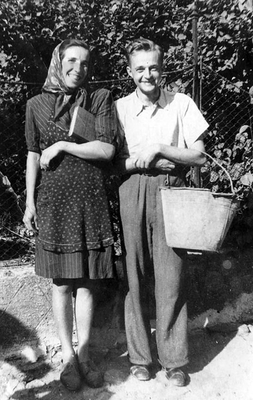 Jan Bula as a young man in a white shirt and dark trousers, a pail in his hand, standing beside a smiling woman in a headscarf at a wire fence
