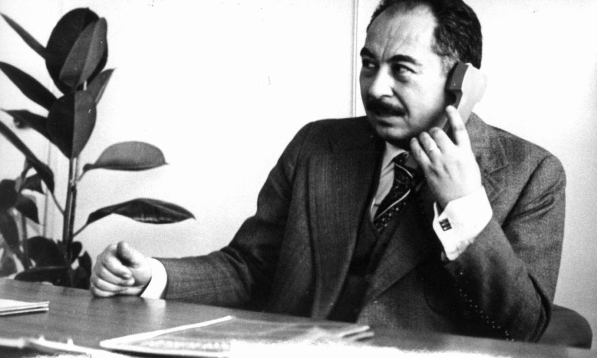 Stjepan Đureković at an office desk in a dark suit and patterned tie, a telephone handset held to his ear, a rubber plant behind him
