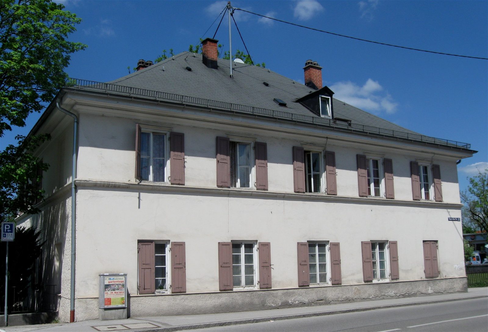 A large white two storey Bavarian house with brown shutters and a hipped grey roof standing directly on Sauerlacher Straße in Wolfratshausen, the street sign for number 15 on its corner