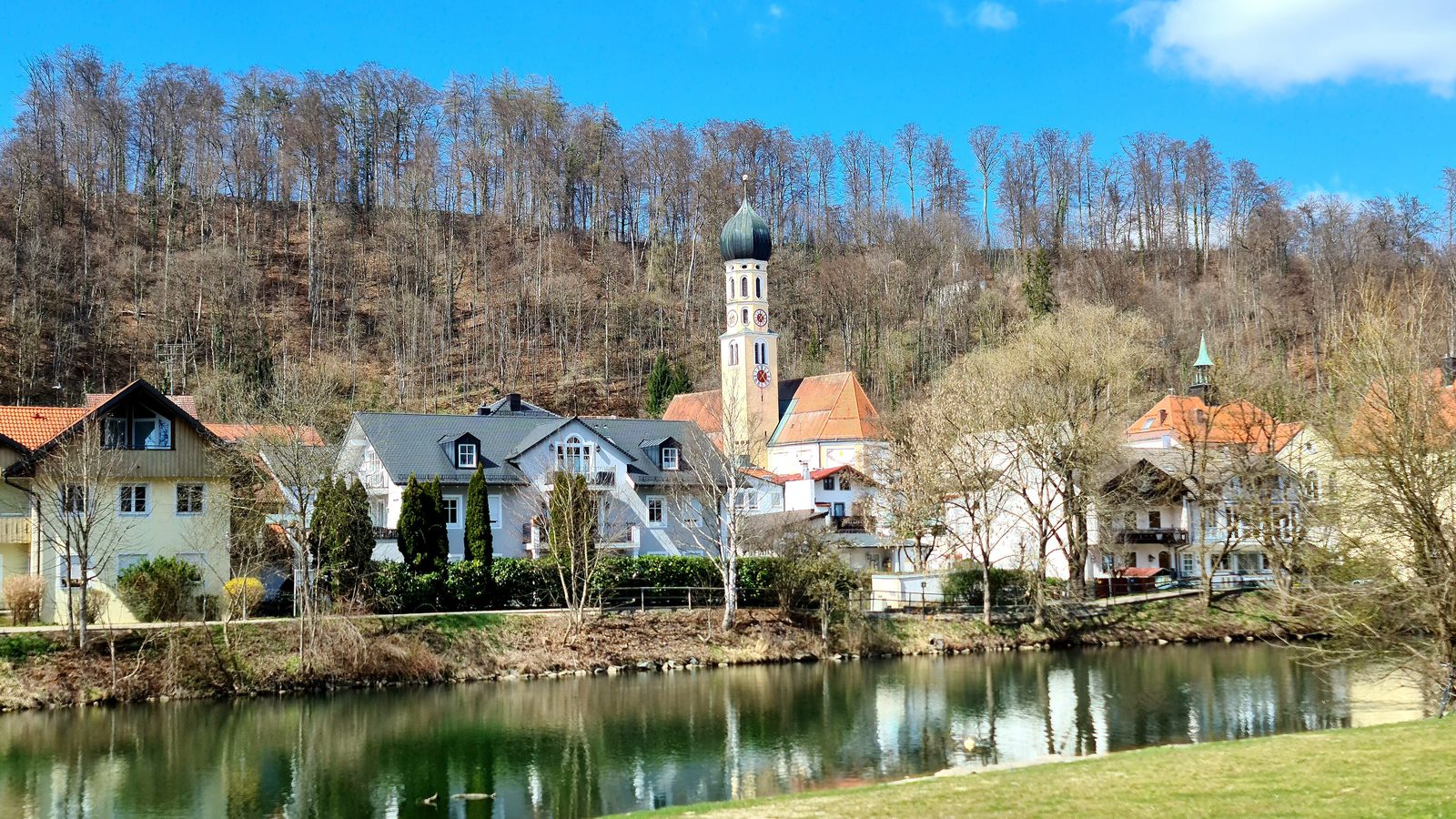 The old town of Wolfratshausen seen from the far bank of the Loisach, painted houses and gardens along the water and the onion domed tower of the parish church against a wooded hill