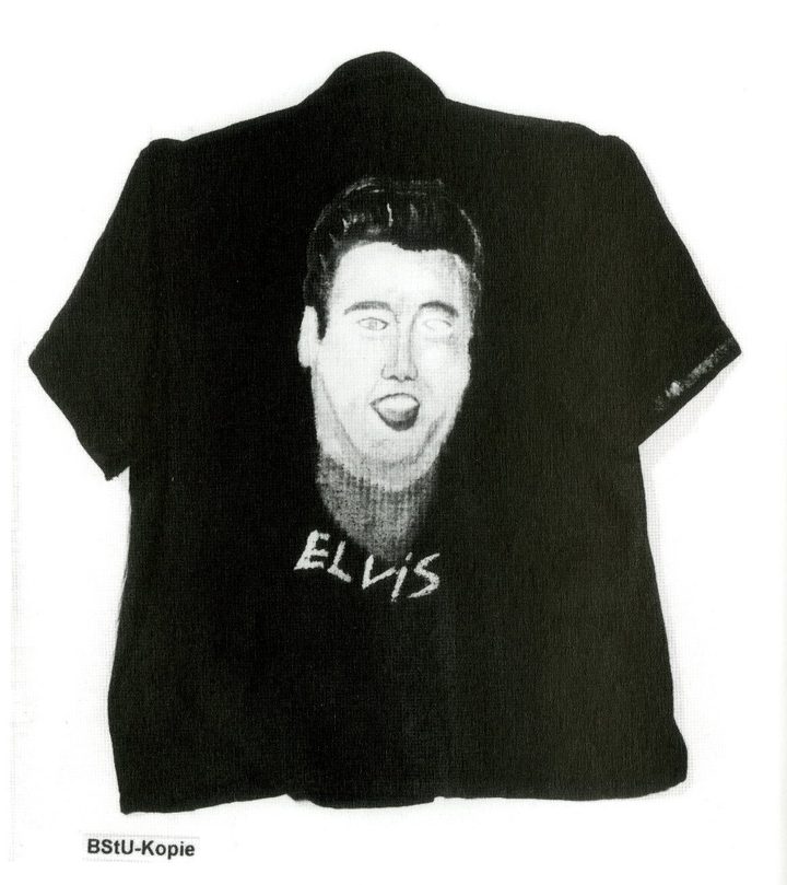 A dark short-sleeved shirt hand-painted with a face and the word ELVIS, on a white ground, with a BStU-Kopie tag