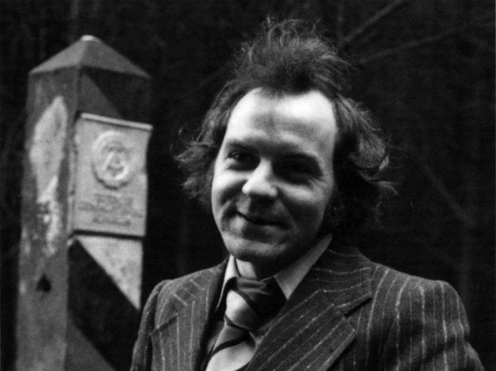 Michael Gartenschläger, smiling, in a pinstriped suit and striped tie, holding binoculars beside a striped East German border post with the state emblem