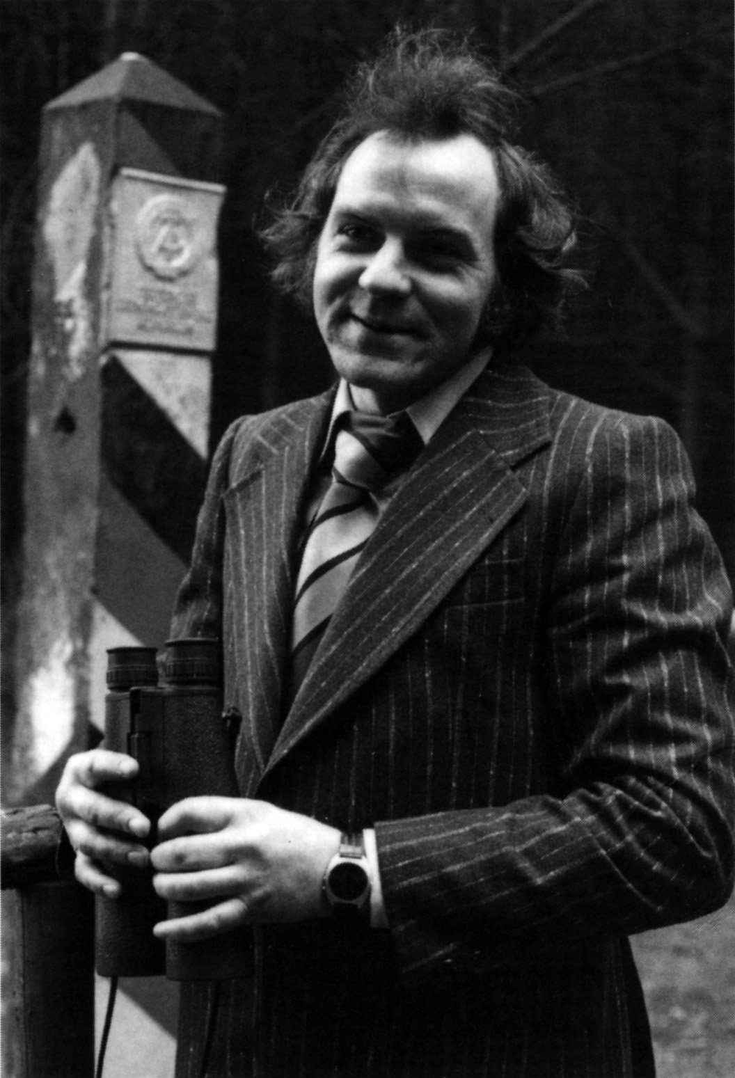 Michael Gartenschläger, smiling, in a pinstriped suit and striped tie, holding binoculars beside a striped East German border post with the state emblem