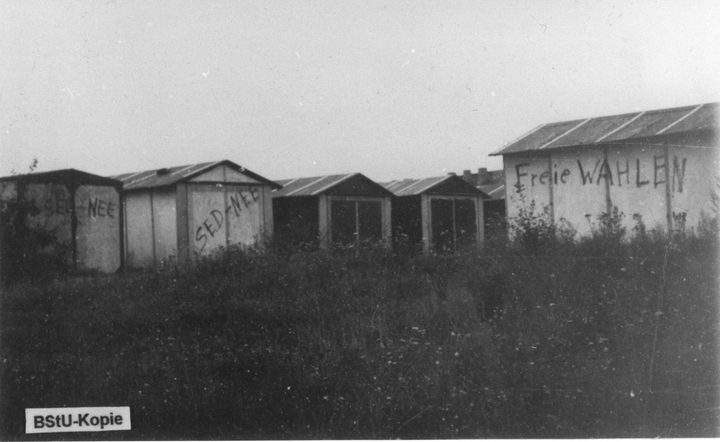 A row of garages and a shed in tall grass, painted with SED NEE and Freie WAHLEN; a BStU-Kopie tag in the corner