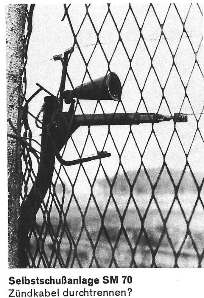 A black and white photograph of a funnel-shaped device fixed to a fence of diamond mesh; captioned Selbstschußanlage SM 70, Zündkabel durchtrennen?