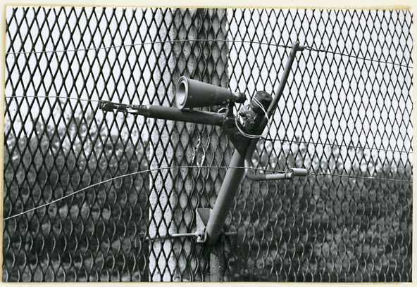 A black and white photograph of a funnel-shaped device on a support tube behind wire mesh, trip wires running from it along the fence