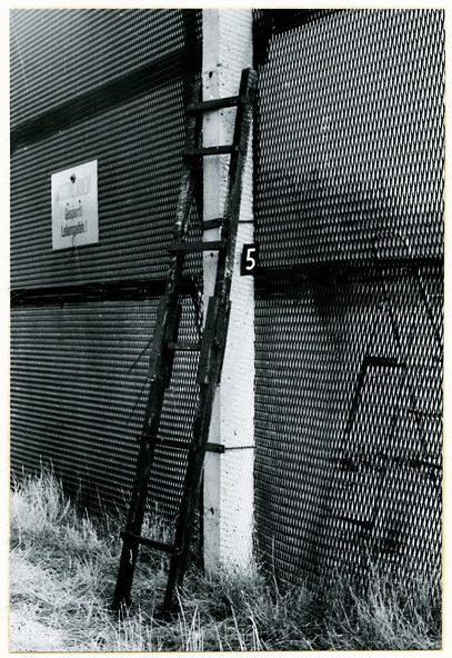 A black and white photograph of a wooden slat ladder leaning on a concrete fence post, a sign reading Achtung Minen! Gesperrt! Lebensgefahr! on the mesh to the left