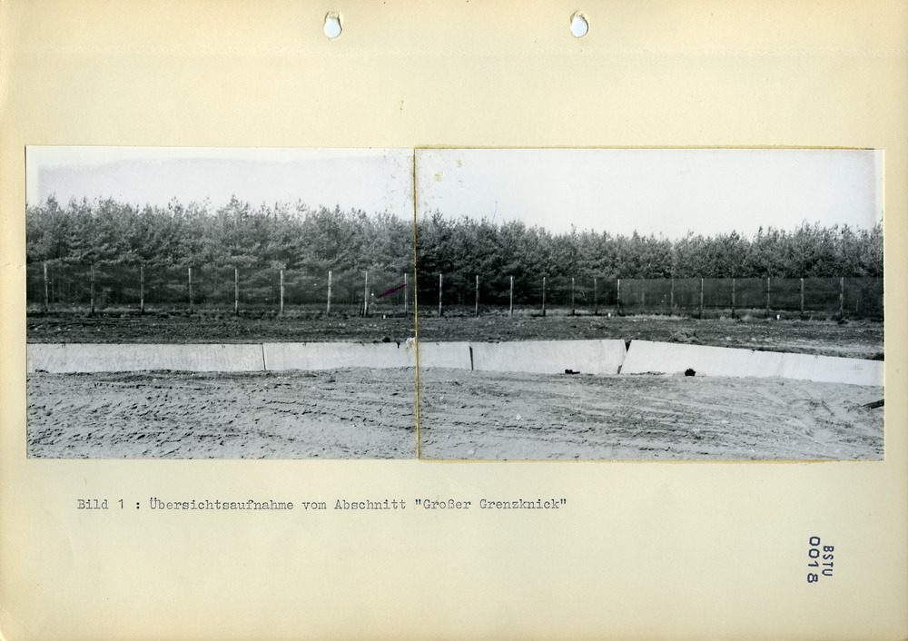 A typed page with two mounted photographs joined into a panorama of the fence line before a pine wood, a red arrow at one post, captioned Bild 1: Übersichtsaufnahme vom Abschnitt Großer Grenzknick