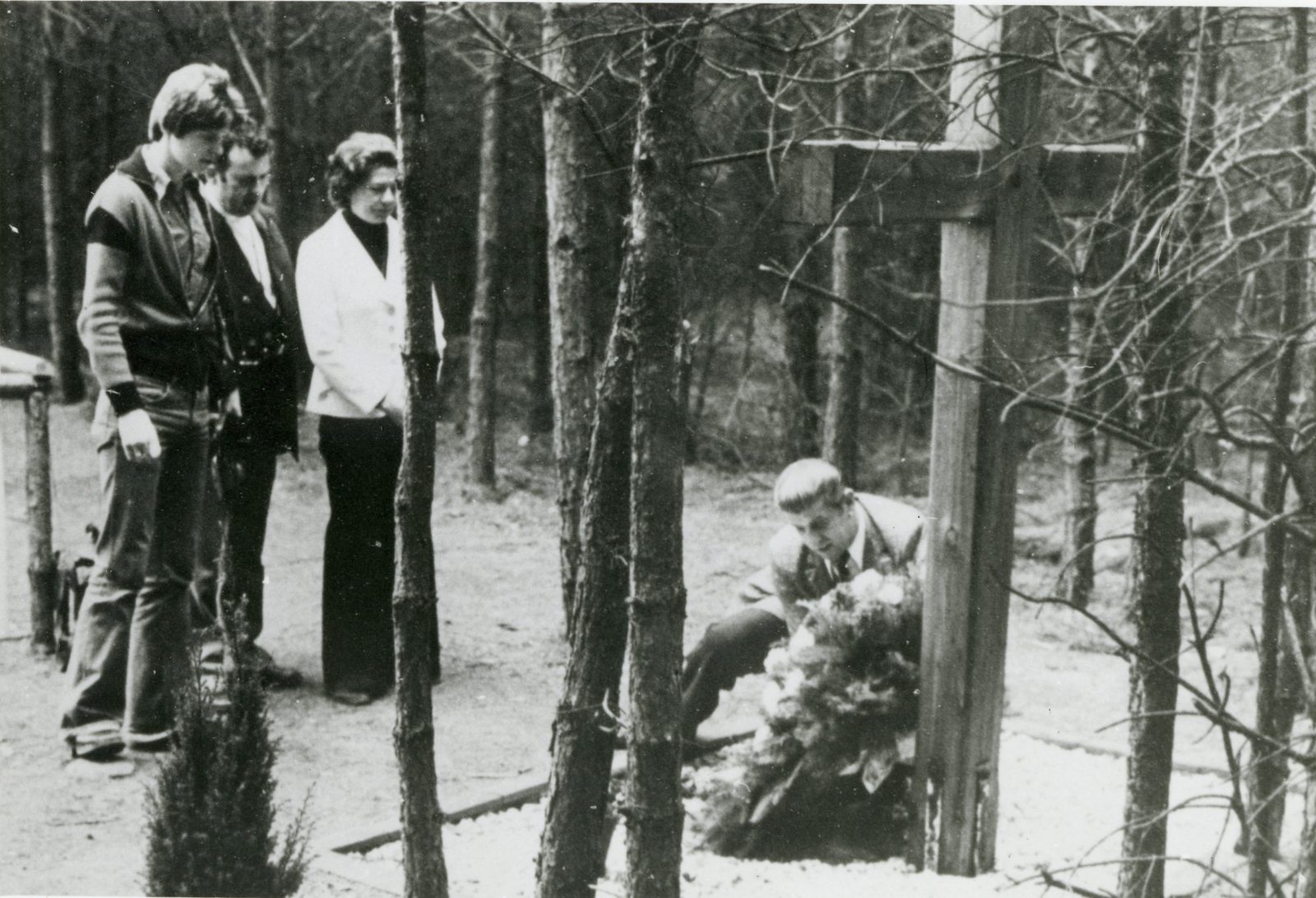 Three people stand among pine trees while a young man lays a wreath at the foot of a wooden cross