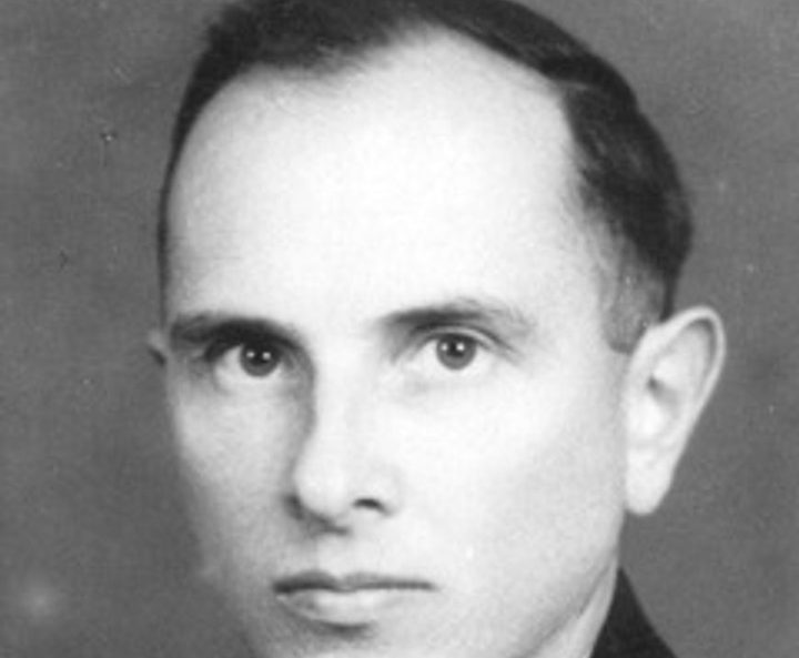 A black and white portrait of a man with receding hair in a dark jacket, a white shirt and a patterned tie