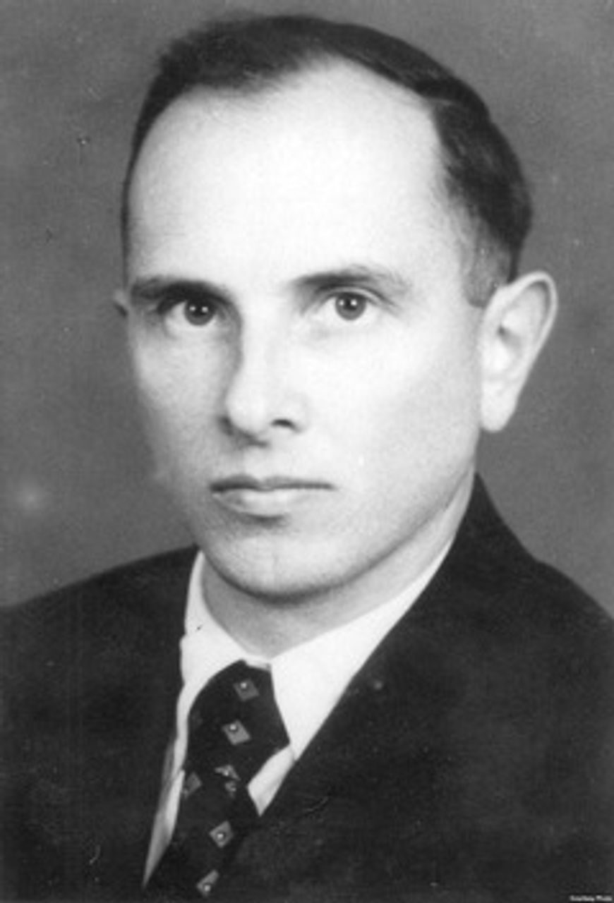 A black and white portrait of a man with receding hair in a dark jacket, a white shirt and a patterned tie