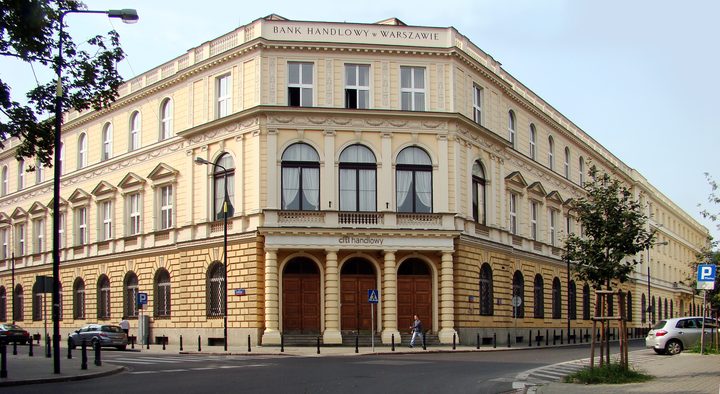 A colour photograph of a cream-coloured corner building with arched ground-floor windows under the sign Bank Handlowy w Warszawie, modern cars parked in the street