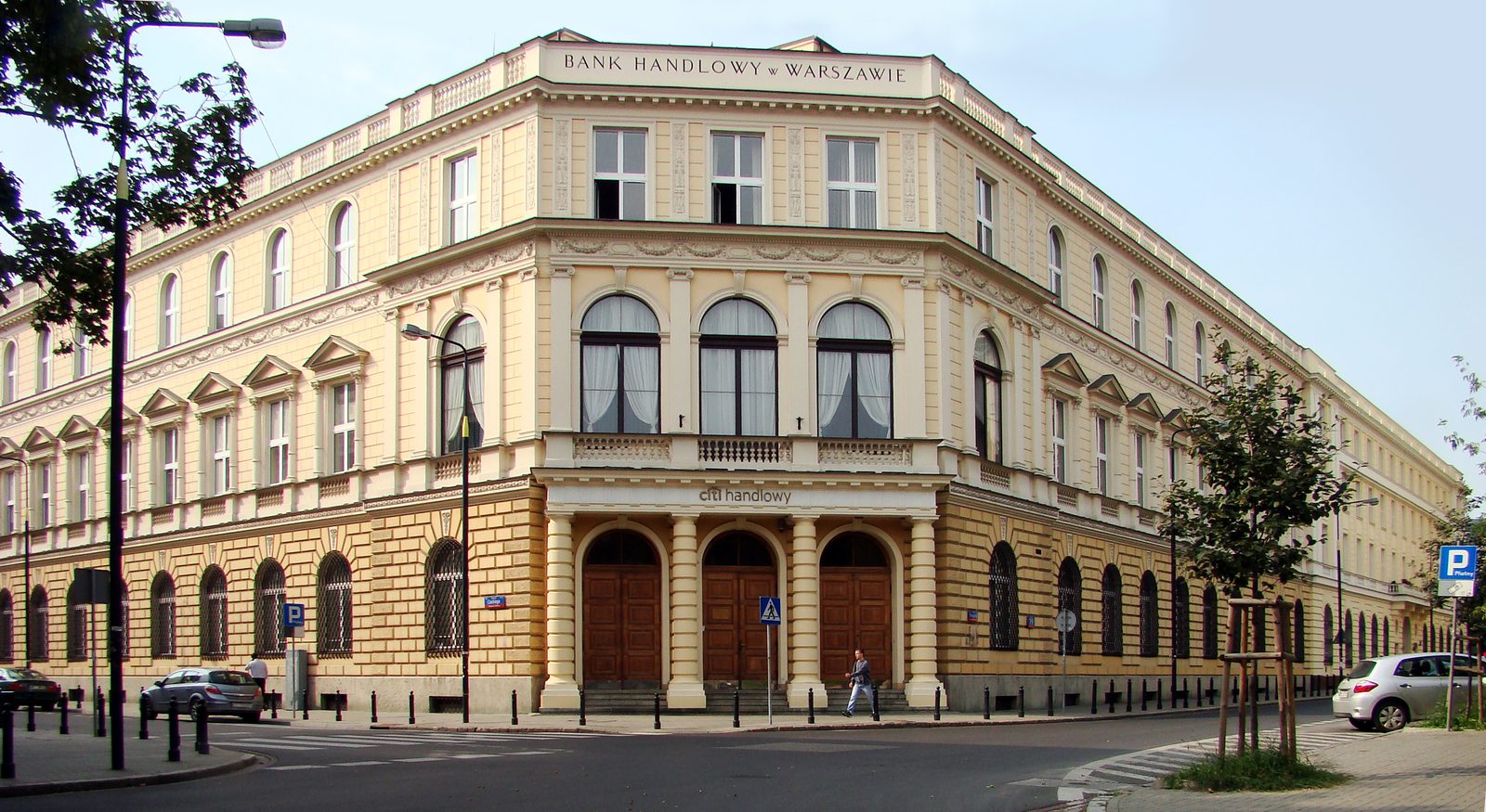 A colour photograph of a cream-coloured corner building with arched ground-floor windows under the sign Bank Handlowy w Warszawie, modern cars parked in the street