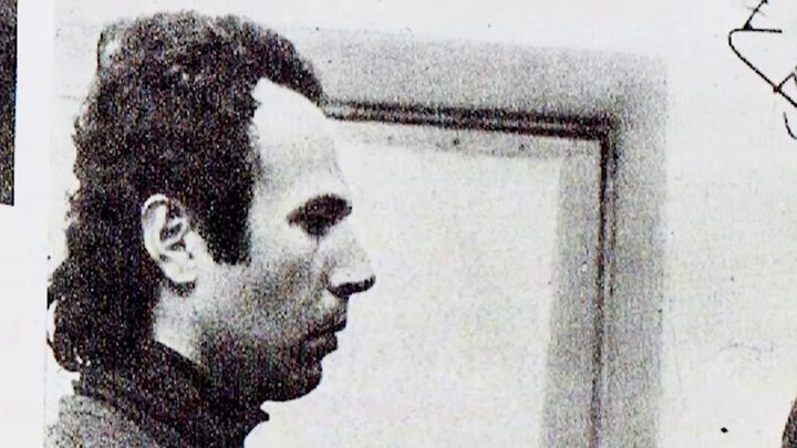 A black and white photograph of a young man with dark wavy hair and a high-necked dark sweater, seen in profile against a pale wall