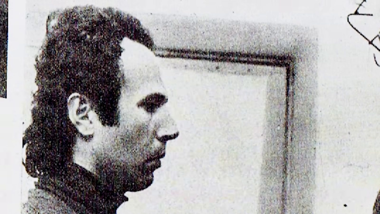 A black and white photograph of a young man with dark wavy hair and a high-necked dark sweater, seen in profile against a pale wall