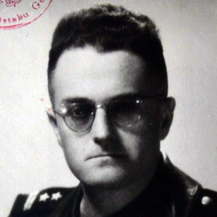 A black and white portrait of a young officer with round glasses and short dark hair, in uniform; a red stamp shows at the top left