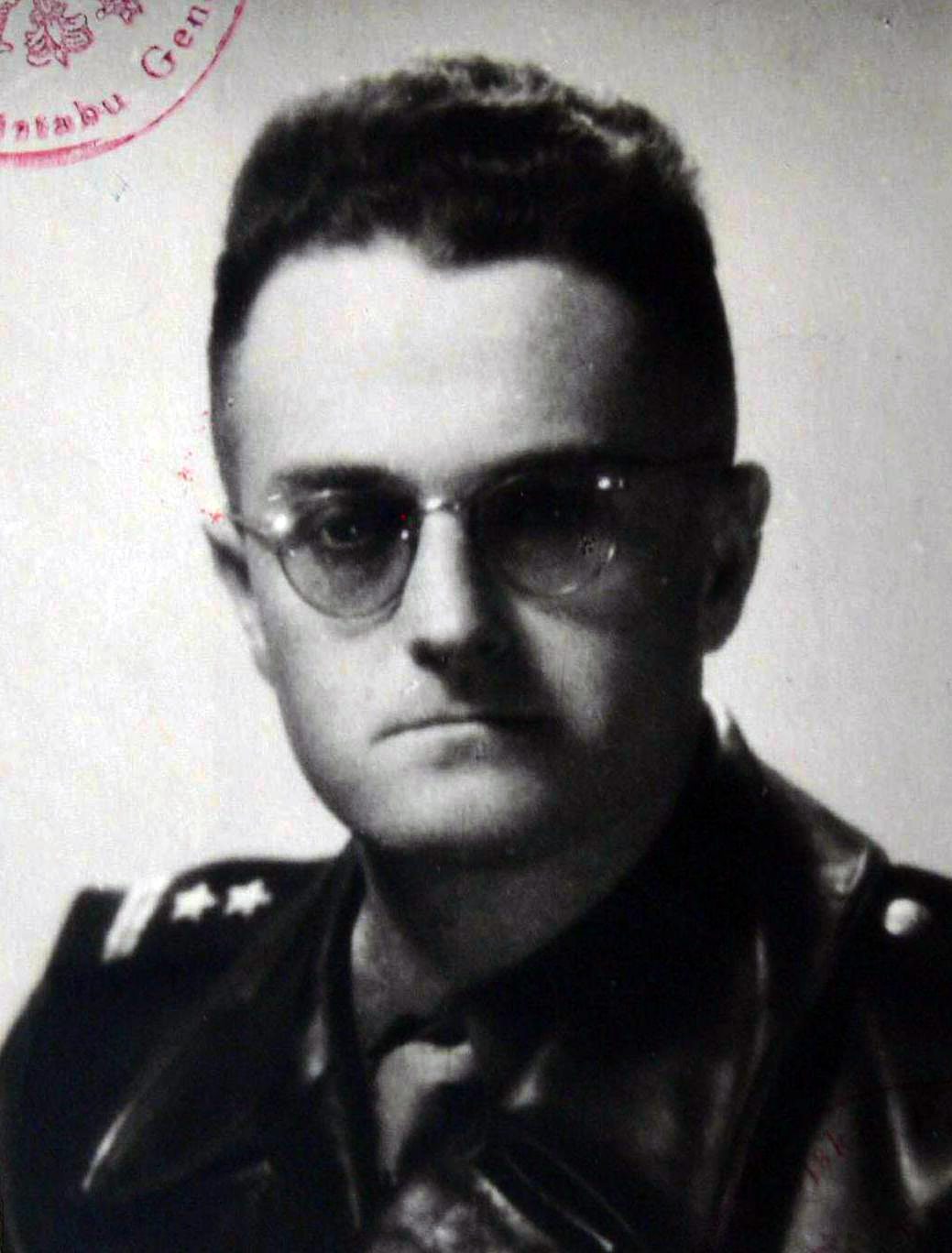A black and white portrait of a young officer with round glasses and short dark hair, in uniform; a red stamp shows at the top left
