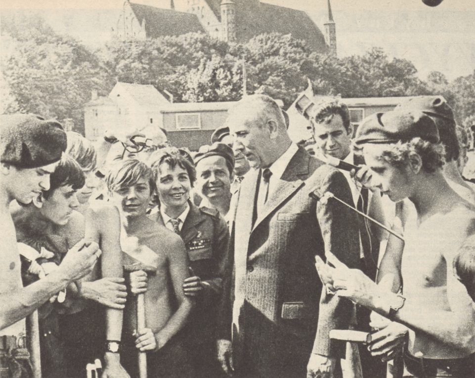 A black and white photograph of a man in a suit among young volunteers, some of them shirtless and in berets, with a cathedral behind them