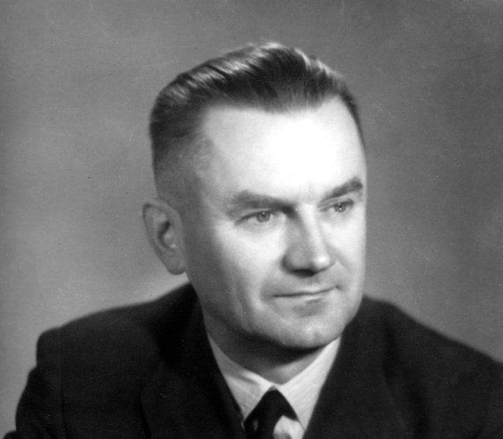 A black and white portrait of a man with short hair, his arms folded, in a dark suit and tie