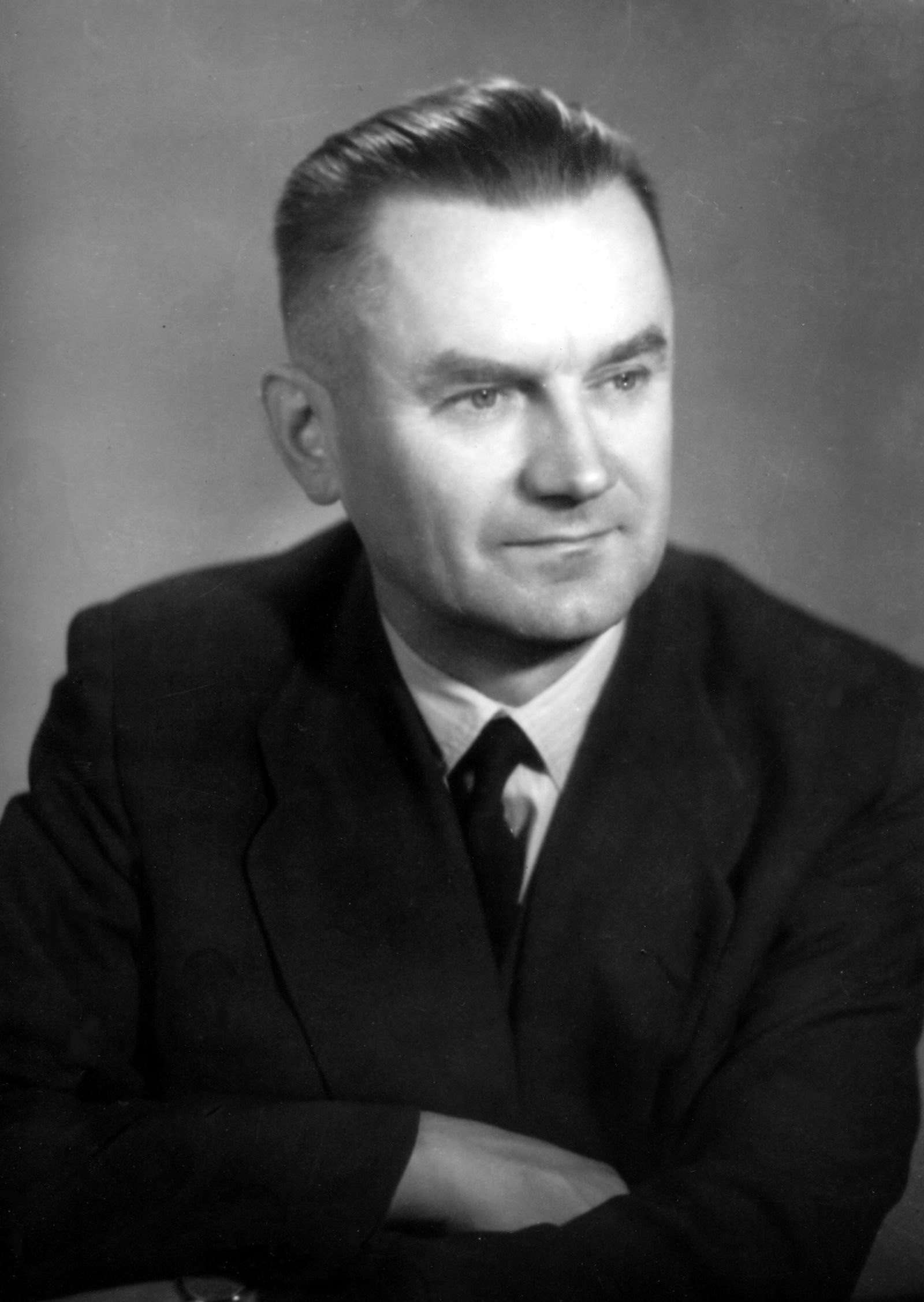 A black and white portrait of a man with short hair, his arms folded, in a dark suit and tie