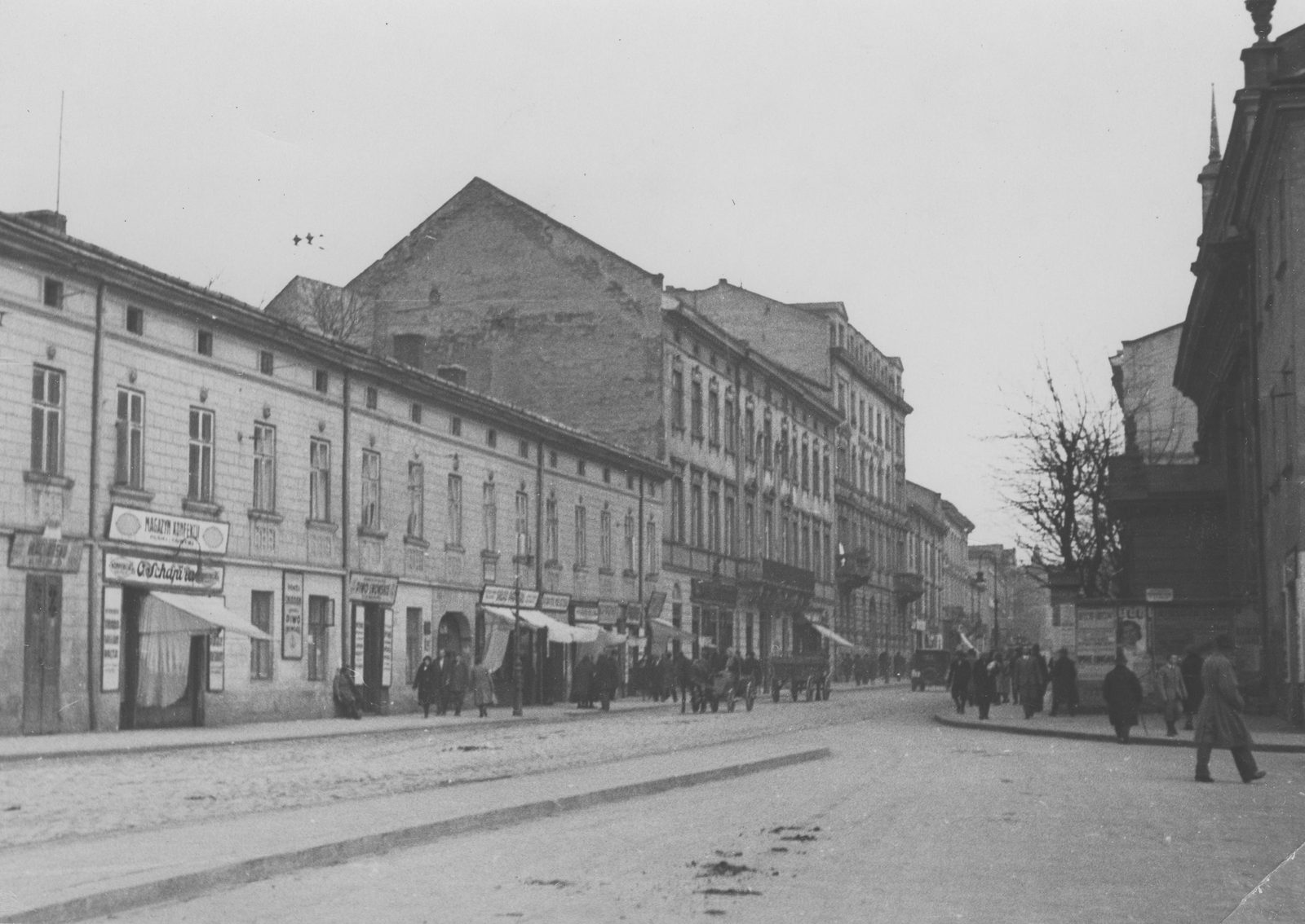 A black and white photograph of a cobbled street lined with two and three storey houses and shop signs, a few people walking