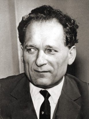 A black and white portrait of a man with thick dark hair looking to the side, in a jacket and dark tie