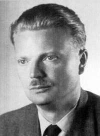 A black and white portrait of a man with combed-back hair and a small moustache, in a jacket and tie