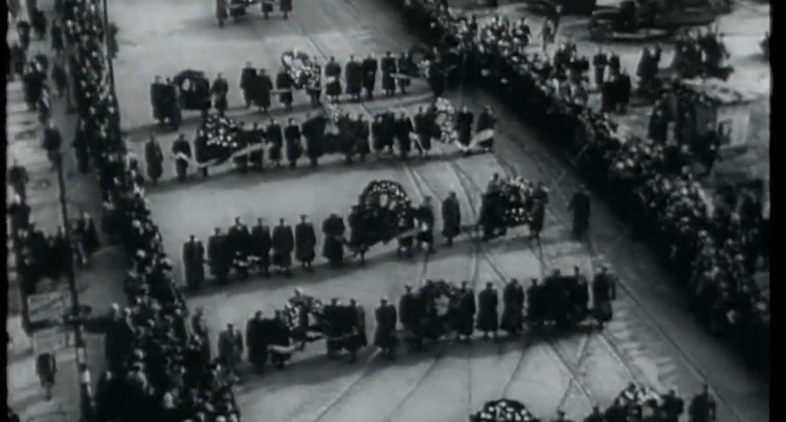 A black and white newsreel view from above of a funeral procession moving down a wide street lined with crowds, wreaths carried in rows