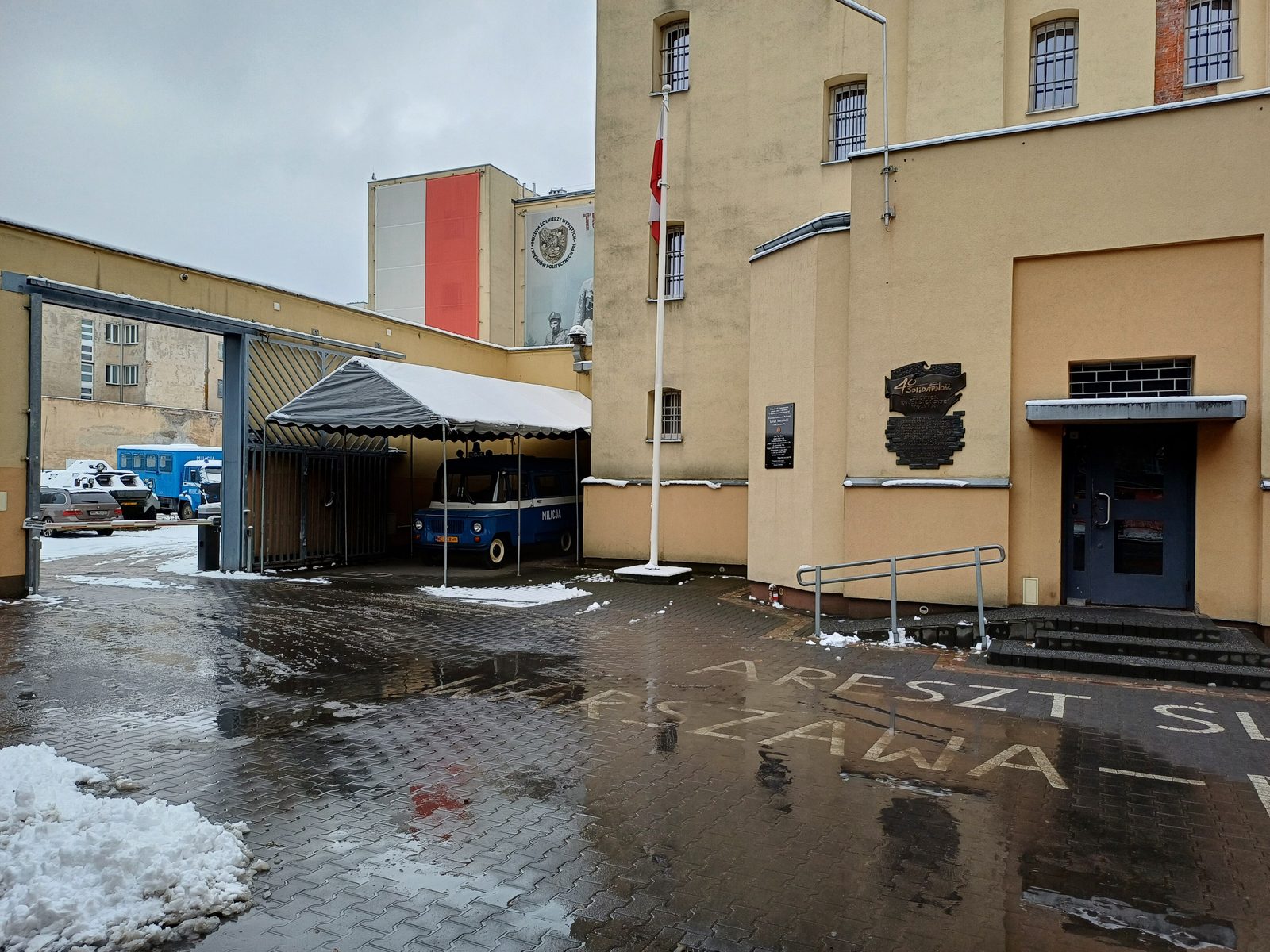 A colour photograph of a prison yard in light snow, a yellow building with a steel door, a flagpole and a parked prison van
