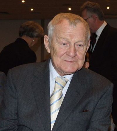 A colour photograph of an elderly man with white hair in a grey suit and striped tie, people behind him