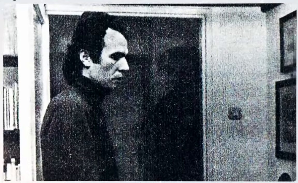 A black and white photograph of a young man in a dark turtleneck, in profile, standing in a doorway; framed pictures hang on the wall to the right