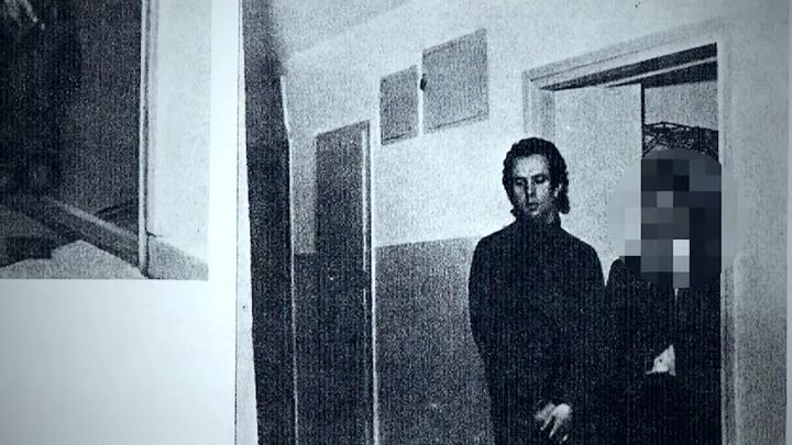 A black and white photograph of a young man in a dark sweater standing in the doorway of a stairwell landing, a second person beside him pixelated