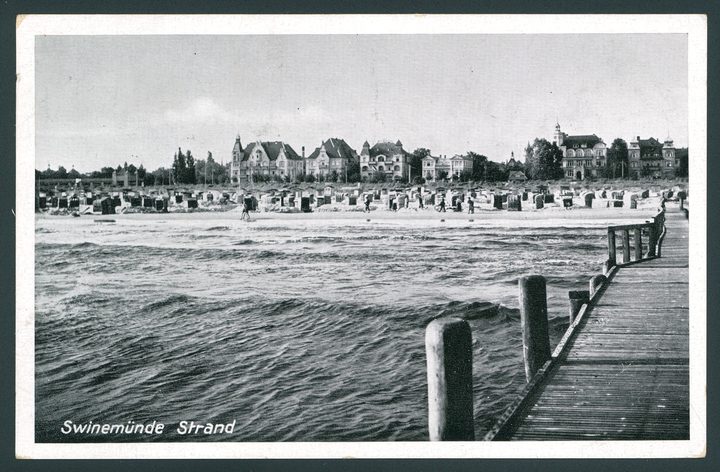 A black and white postcard of a beach with wicker beach chairs and seafront villas, a wooden pier in the foreground, captioned Swinemünde Strand