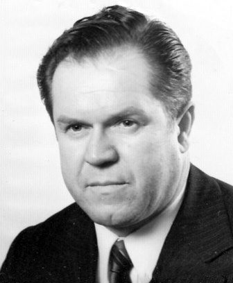 A black and white portrait of a heavy-set man with swept-back hair in a dark suit and tie