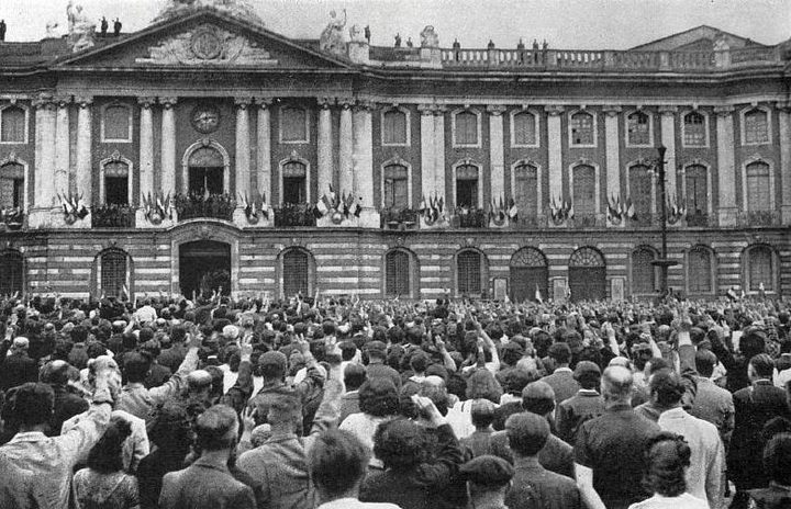 A black and white photograph of a large crowd filling a square in front of a long classical facade hung with flags