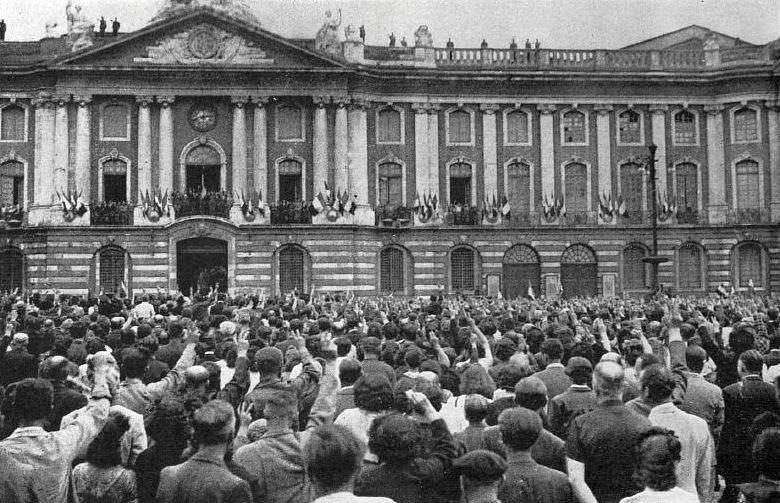 A black and white photograph of a large crowd filling a square in front of a long classical facade hung with flags