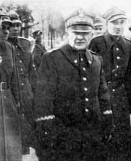 A black and white photograph of an older general in a peaked cap and greatcoat among officers in caps