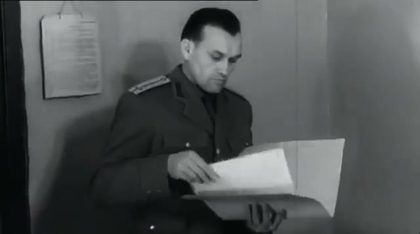 A Securitate officer in uniform, seated, reading an open file held on his knee, a printed notice pinned to the wall behind him