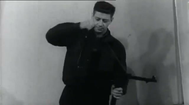 A man in a dark jacket pulls a garment over his head, a submachine gun in his other hand, standing against a bare wall