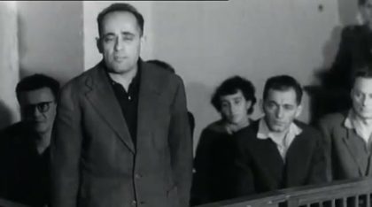 A man stands at the front of a wooden dock, hands at his sides, with five others seated behind him, one of them a woman and one wearing glasses, filmed in black and white