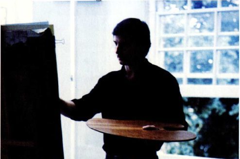 A colour photograph of a man standing at an easel by a window, a wooden palette in one hand