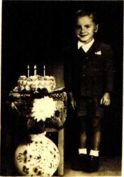 A sepia photograph of a small boy in a jacket and shorts standing beside a decorated birthday cake with lit candles on a stand