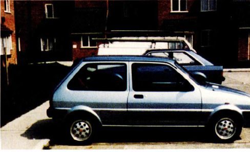 A colour photograph of a small silver hatchback car parked on a paved forecourt in front of a low brick block of flats, a second car behind it