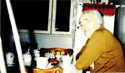 A colour photograph of an elderly man with white hair turning towards the camera at a table laid with cups and fruit, in a room with a curtained window