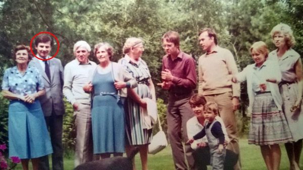 A colour photograph of a family group of about a dozen adults and two children standing on a lawn in front of trees; the head of a dark haired man in a suit, second from the left, is ringed in red