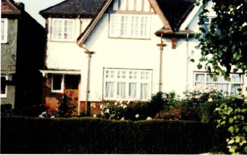 A colour photograph of a white rendered semi detached suburban house with bay windows and a mock timbered gable, behind a clipped hedge and a rose bed